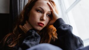 Mujer joven pensativa junto a una ventana, con la mano en la frente, preocupada