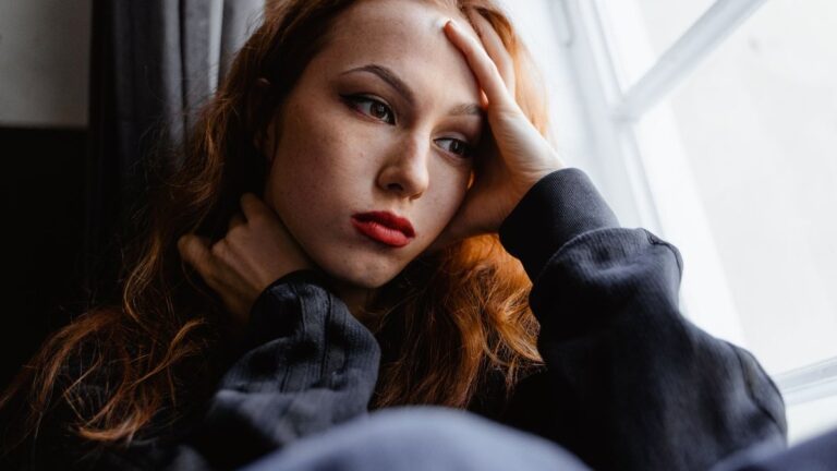 Mujer joven pensativa junto a una ventana, con la mano en la frente, preocupada